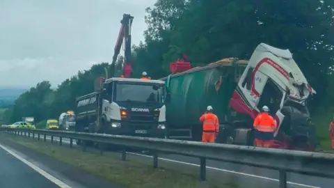 Liz Kirkhope A lorry with its cabin crushed following a collision. Men in hi-vis stand beside the lorry and another vehicle with a winch