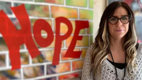 Alex Pope/BBC Hayley Brown standing by a Hope sign, in red, painted to look like a wall, Hayley is wearing a cream jumper, long necklace, with a black top underneath. She has long dark hair and is wearing glasses.