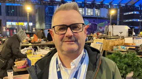 A man stands smiling in front of market stalls. He has black rimmed glasses, a green coat and a white shirt with a lanyard.