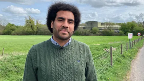 A man with Afro hair and a close shaven beard is looking at the camera with a field behind him. He is wearing a green cable-knit jumper and blue shirt.