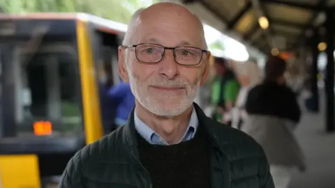 Bill Dodds smiling into the camera. He is mostly bald with white hair by his temples. He is wearing dark, rectangular glasses and a black coat and jumper with a light blue shirt underneath. He is standing in front of a black and yellow old Metro train which is out of focus.