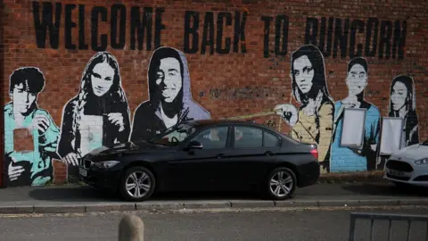 A painting of teenagers on a brick wall under writing which reads "Welcome Back To Runcorn". A black car stands in front of the wall