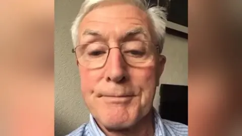 John Richards A man with silver hair and glasses looks down while in front of a webcam, with his face and neck visible