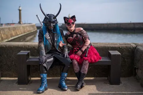 Getty Images Two costumed people sit on a wooden bench by a coastal harbor. A lighthouse stands in the distance to the left. Both people are wearing elaborate, theatrical outfits that resemble fantasy or gothic-inspired costumes. The person on the left is dressed in a black leather outfit that includes a long leather coat, fitted trousers, and black gloves. A katana-style sword in a white sheath is strapped diagonally across their back. The person on the right is wearing a red-and-black dress with a fitted bodice and a layered, ruffled red skirt that flares outward.