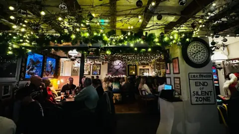 St Mabyn Inn Dozens of people sit inside the St Mabyn Inn in Cornwall. Fairy lights are on the roof. The pub is dimly lit.