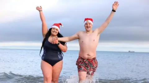 PA Media Rebecca, left, is wearing a navy swimming costume and red and white Santa hat, she has her arm up in the air. She is holding hands with Joel, right, who has patterned swimming shorts n and a red and white Santa hat.