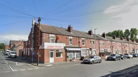 A row of terraced buildings with cars parked on the roadside. There is a junction to the side leading on to another road. One of the buildings has a white sign outside with orange writing which reads 'Jumbo House'.