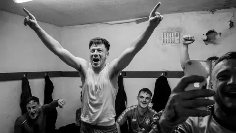 John Bolloten A black and white photo of a man inside a changing room cheering with both his hands raised in the air. Others behind him are also in good spirits.
