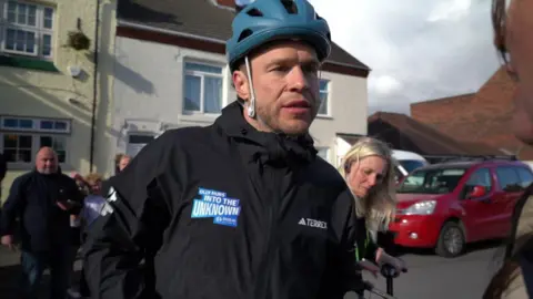 BBC A man wearing a blue cycle helmet and black jacket stands in the street. The jacket is branded "Olly Murs into the Unknown". A small crowd of spectators stands behind him on his route.