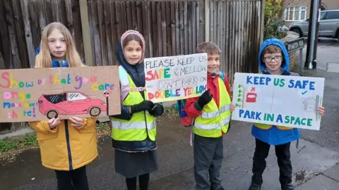 Becky Carlyle Four children wearing coats and hi-vis vests stand on the pavement holding signs. One reads: "Show us that you care or go move over there." Another says: "Please keep us safe and mellow. Don't park on double yellow." The third sign says: "Keep us safe. Park in a space."