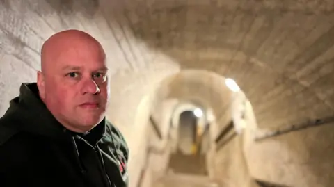 Malcolm Amy stood at the entrance to an underground tunnel. There are steps going down and the brick is white. Malcolm Amy is stood on the left and is facing the camera, he is wearing a green hoodie.