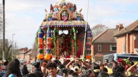 Matt Turpin A brightly coloured Hindu float is paraded through a street. Terraced houses can be seen in the background and a crowd of people are surrounding the float.