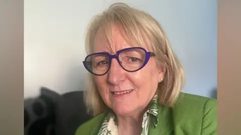 Jones has short blonde hair and a fringe. She is wearing purple glasses, a green blazer and patterned shirt.