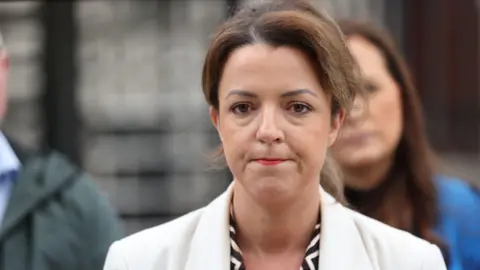 PA Media Infrastructure Minister Liz Kimmins speaking to the media outside Belfast High Court she is wearing a white blazer and black and white shirt. She has brown hair