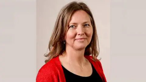 Mandy Clare is smiling. She has long brown hair and is wearing a black top and red cardigan.