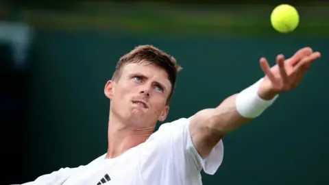 Harris, serving at Wimbledon - his arm is outstretched and his eyes are fixed on the yellow ball in the air. He is wearing white and has short brown hair.