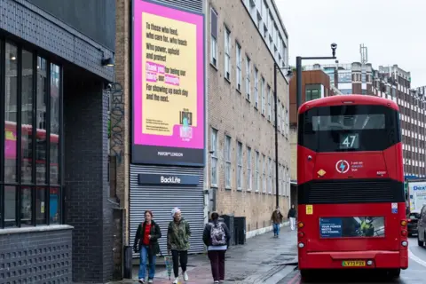London City Hall A number 47 bus drives past a pink and yellow sign from the campaign. Three people walk along a pavement beneath the sign