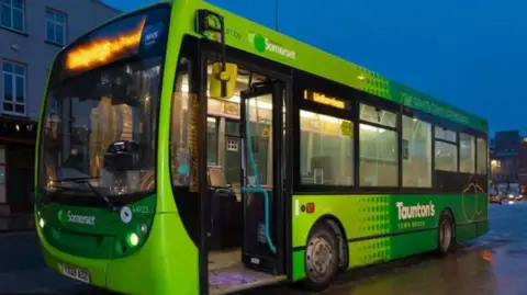 A green bus with Somerset written on the side and the front. The bus is stationary and door is open. You can see a road in the background.