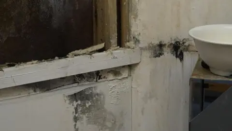 A wall inside the school has paint flaking off it and there are black marks and pieces of plasterboard.