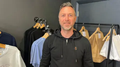 Man with grey hair and black hoodie stands infront of wall of t shirts 