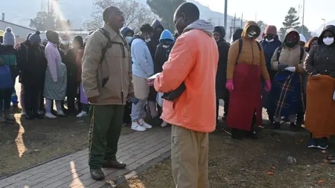 Khanyisile Ngcobo / BBC is a man in an orange jacket carrying a prayer book leading improvised service in public places. Another man stands in front of him with his eyes closed in focus.