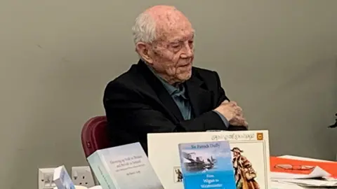 BBC Sir Patrick Duffy sitting at a table behind a display of his books and artwork, there are papers and a pair of glasses on the table and he is wearing a turquoise polo shirt and a navy-coloured blazer.