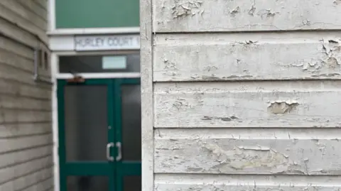 A section of wooden panelled wall is in the foreground - it's painted white, but the paint is peeling away and looks slightly grey and weathered. Blurred in the background is the entrance to Hurley Court. A double door, which is a dark green with glass panels can be seen, with a white sign above which reads 'Hurley Court'