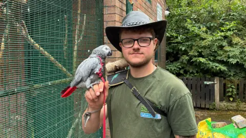 A picure of Connor Atherton who is wearing a hat and green t-shirt and black glasses. He smiles into the camera, whilst holding a gray parrot. 