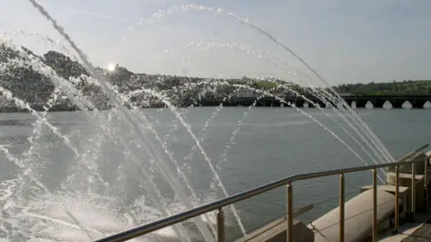A water fountain spraying water into a river. In the background is a bridge. There is a hill with houses. The sky is a light blue with the sun coming out from behind a house. The river is a dark blue. The water coming out of the fountain is clear. 