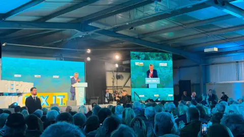 A crowd of people listening to a man in a dark blue suit delivering a speech on stage. The conference room is illuminated with teal-coloured lighting. 