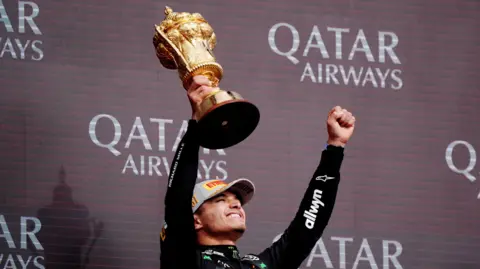 Lando Norris smiles and lifts a large gold trophy in the air in front of a board that advertises Qatar Airways. He lifts the trophy in his left hand and raises his right fist in the air