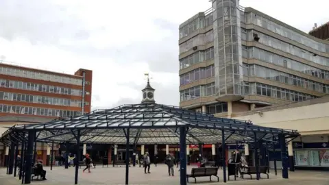 People walking through a large gazebo with blue columns and a clock tower. Two multi-storey buildings are either side of the gazebo