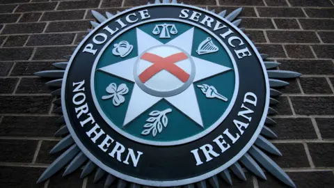 A PSNI crest on a brown brick wall. The crest is a circular white, green and black badge, outlined by a silver star. 