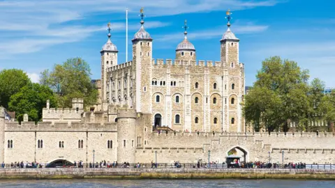 The Tower of London viewed from across the River Thames, showing its stone walls, corner towers with domed roofs, and surrounding grounds under a blue sky, with people walking along the riverside.