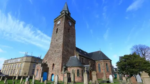 The picture has been taken looking up at the church and its clock tower. There are gravestones in the foreground and blue skies with wispy cloud above.