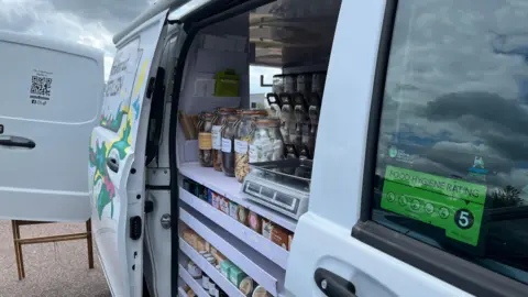 Tom Jackson/BBC A white van with its door open. Inside, there are small shelves containing items such as soap and shampoo and above them is a counter with a weighing scale and jars of dried food. 