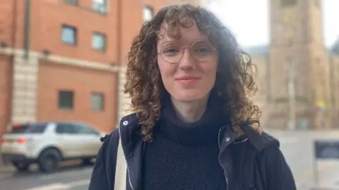 Ash Black wears a black jumper and jacket. She has curly brown hair and glasses. Behind her is a car and buildings. 