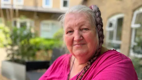 Head and shoulders image of Harmony Birch. Harmony has her hair in a plait to the side, is wearing a pink top and sitting in a garden.