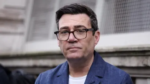EPA Andy Burnham, wearing black frame glasses and a blue jacket with a white T-shirt, looks at the camera