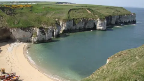 Caroline Thorogood A landscape shot at the top of grassy and white cliffs overlooking a beach area and sea. Two small boats can be seen on the sand.