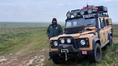 Man standing outdoors alongside mustard-coloured Range Rover with camping equipment attached. It is parked grass in a rural area. He has on a dark green ambulance jacket and a black beanie hat, and has a large, dark beard.