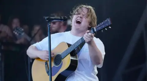 PA Media Lewis Capaldi performs on stage, while holding a guitar.