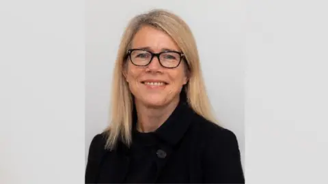 A woman wearing a black coat and black glasses. She is smiling and has shoulder length blonde hair. Behind her is a grey background.