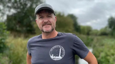 BBC/Jo Makel A man with a greying beard stands on the banks of the River Freshney in Grimsby. He is wearing a t-shirt and a baseball cap.