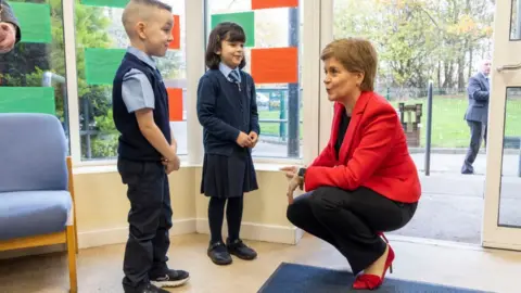 Getty Images Nicola Sturgeon, with short fair hair, crouches in front of two primary school children. She is wearing a red jacket and high heels with black trousers. They are in a primary school. 