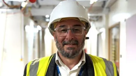 Qays Najm/BBC Stephen Balls with beard and glasses wears a hard hat and high-visibility vest. He stands in a hospital corridor where construction work is being carried out.