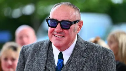 A man with short grey hair wearing large black glasses with blue tinted lenses. He is wearing a grey suit, white shirt and a blue tartan tie