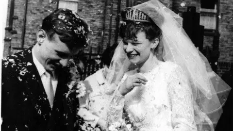 Vin Rigby Black and white photo of a happy bride and groom in traditional wedding outfits. They laugh as confetti falls on the grooms hair and the bride's white dress.