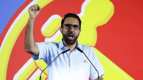 Getty Images Singapore's Workers' Party leader Pritam Singh speaks at a rally, standing on a podium where he pumps his fist in the air. In the background is a large red and yellow sign of the party's hammer symbol