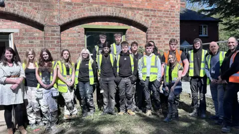 BBC/Kate Bradbrook Fourteen students in hi-vis vests stand alongside teachers, and industry professionals outside the brick cottage. They are all smiling at the camera. 